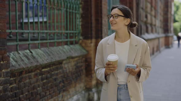 Intelligent Young Woman Walking on Street Holding a Cup of Coffe or Tea and Texting Message on