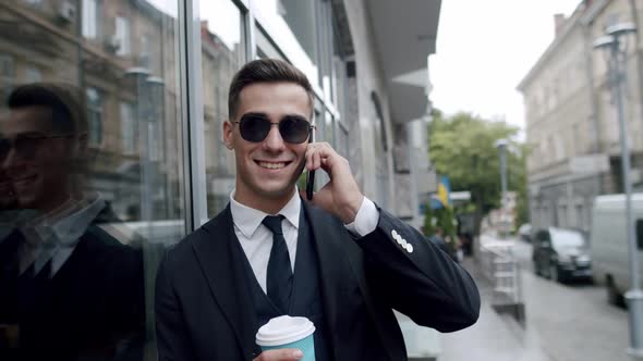 Happy Man Sunglasses Talks on Smartphone and Holds a Cup of Coffee Outdoors