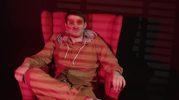 Young man in glasses sitting in armchair in dark room with falling red light and shadow blinds.