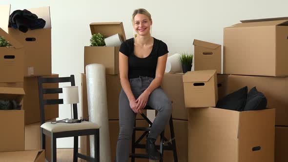 A Happy Moving Woman Sits on a Chair and Smiles at the Camera in an Empty Apartment