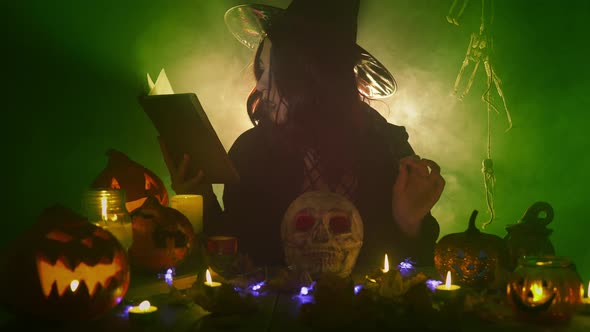 Young Gothic Witch in Dark Mantle and Hat is Conjuring Over the Skull Among Halloween Decorations