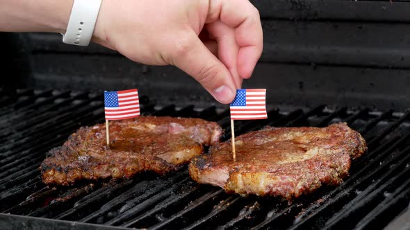 Two juicy rib-eye steaks sitting on the grill and cooking a hand comes in and sticks a tiny American