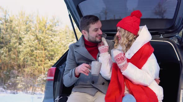 Young Couple a Man and a Woman are Sitting in the Trunk of a Car in a Winter Snowy Forest Hugging