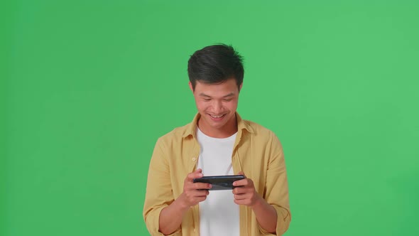 Smiling Asian Man Looking At The Phone Screen And Holding It In His Hands In Green Screen Studio