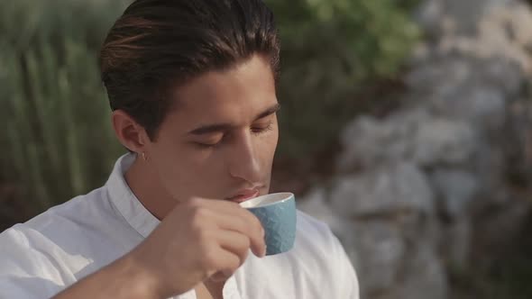 Attractive young fashion man in a suit drinks coffee in the morning outdoors in the garden