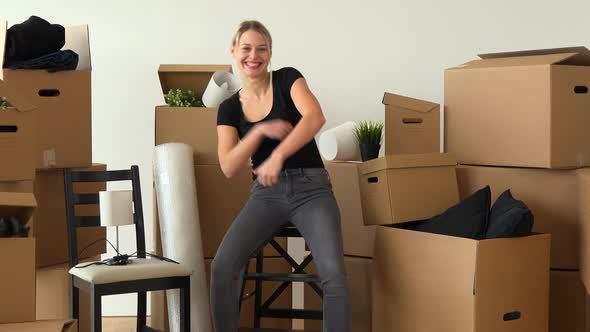 A Happy Moving Woman Sits on a Chair in an Empty Apartment, Dances and Smiles at the Camera