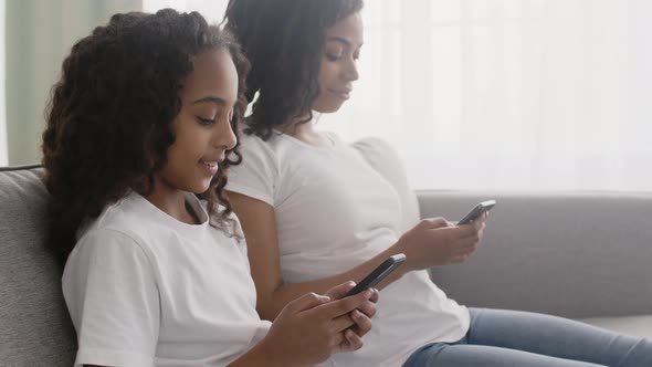 Little African American Girl and Her Young Mother Networking in Smartphones Sitting on Sofa and