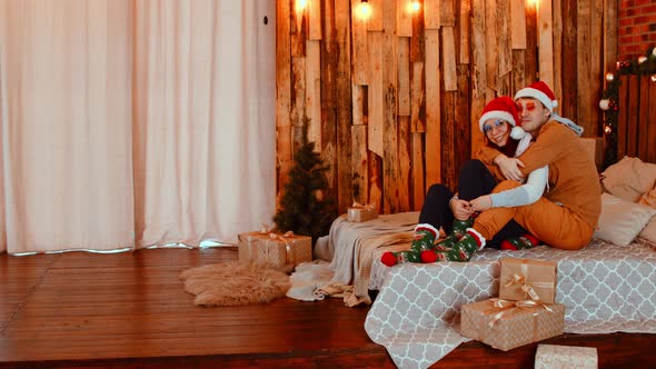 Man and Woman in Santa Hats and Glasses Hugging Tenderly Sitting on Bed