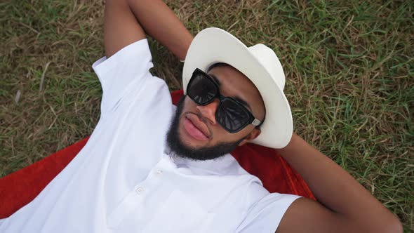 Top View of Carefree Handsome Man in White Hat Polo Shirt and Sunglasses Lying on Green Grass