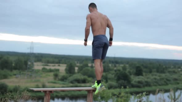 A Young Strong Athlete Without a Shirt Trains on the River and Jumps on the Bench with His Feet in