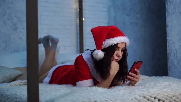 Young Woman in Santa Claus Costume Browses Smartphone Lying on Bed
