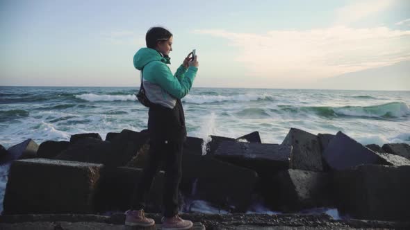 Attractive Young Woman in a Warm Jacket Walking on the Stone Beach at Sunset