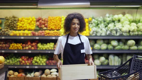 Beautiful Smiling Young Female Supermarket Employee in Black Apron Holding a Box