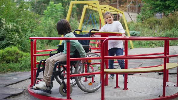 Wide Shot Positive Pretty Girl Spinning Merry Go Round with Disabled Cute Boy in Wheelchair