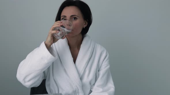 Thirsty smiling 35s woman in white bathrobe drinking water