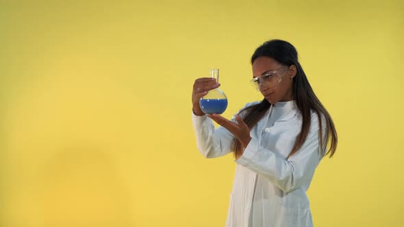 Black Female Scientist in Lab Coat Looking on Flask with Experimental Liquid on Yellow Background.