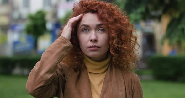 Attractive Red-haired Woman with Blue Eyes Looks Surprised at the Camera Touches Her Face with Her