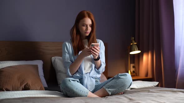 Surprised Redhead Young Woman Getting Good Online News on Mobile Phone at Cozy Bedroom