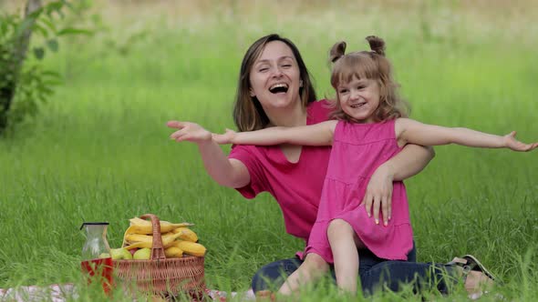 Family Weekend at Picnic. Daughter Child Girl with Mother Sit on Grass Meadow and Waving Hands