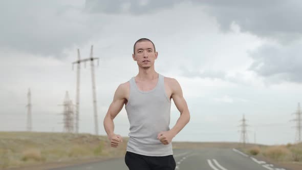 Asian Man Runs Along Road in Sports Clothes Cloudy Weather Front View