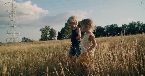 Brother and sister walk on mountain meadow at sunset holding hands, cows grazing