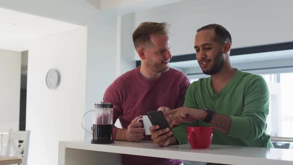 Multi ethnic gay male couple in kitchen talking one using smartphone