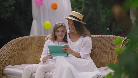 Young and Teenage Sisters Watching Online Film Smiling Sitting on Couch in Spring Summer Garden