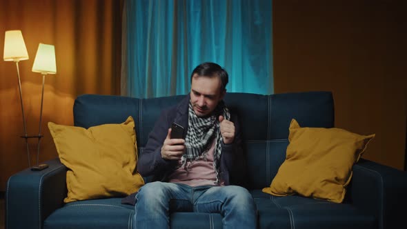 Happy Young Man Sitting on Couch and Looking at Mobile Phone While Singing and Dancing to Music