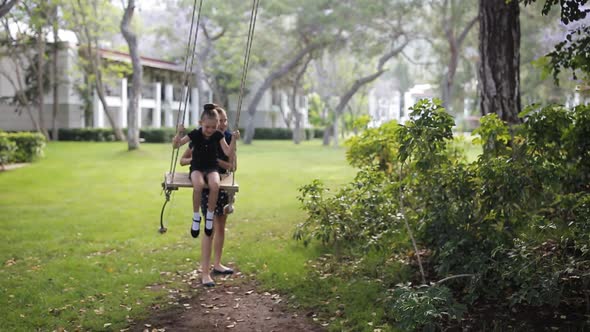 The Older Sister Shakes Her Daughter on a Swing Under a Tree in the Sun