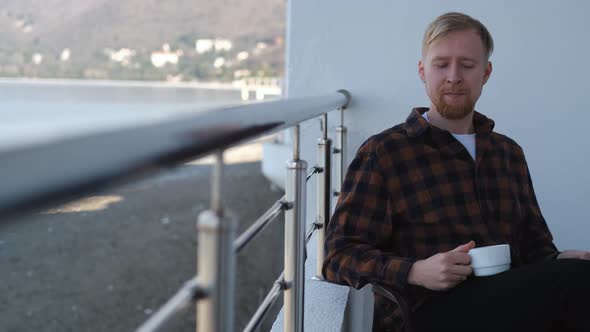 a man with a smartphone sits on a balcony overlooking the sea