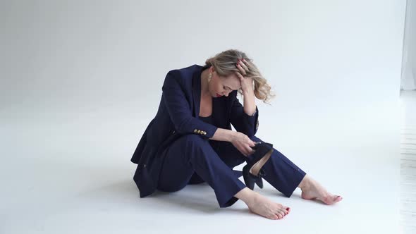 Woman in Blue Suit Sits Barefoot on in White Photo Studio and Holds Heeled Shoes