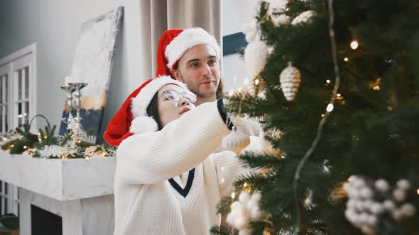 Young Multiracial Couple are Smiling Decorating Christmas Tree with Baubles Standing Nearby a