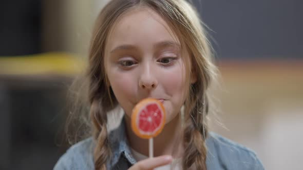 Headshot of Joyful Caucasian Girl with Ponytails Licking Lollypop Looking at Camera and Smiling