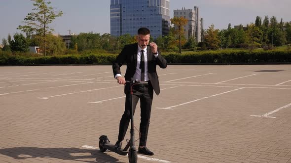 A Businessman Stands with an Electric Scooter in the City, He Puts on Sunglasses