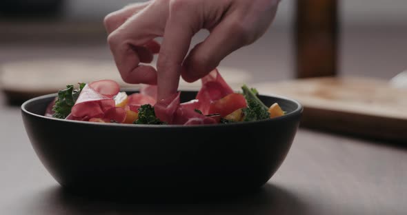 Slow Motion Man Hand Add Coppa Slices To Make Salad with Kale and Cherry Tomatoes in Black Bowl on