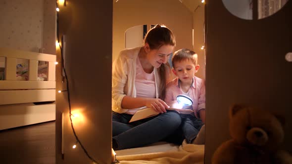 Happy Smiling Boy with Young Mother Sitting in Tent or Toy Cardboard House and Reading Book with