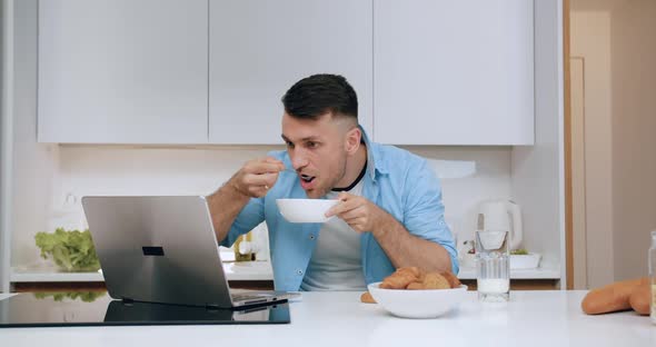 Man Sitting at Kitchen Table, Eating Cereal for Breakfast and Watching Funny Videos on Laptop