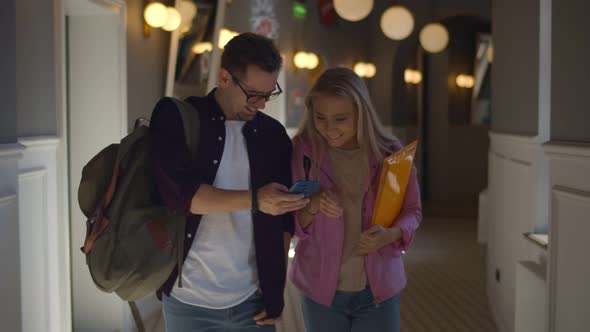 Joyful Student Couple Talking and Using Cellphone While Walking in College Corridor