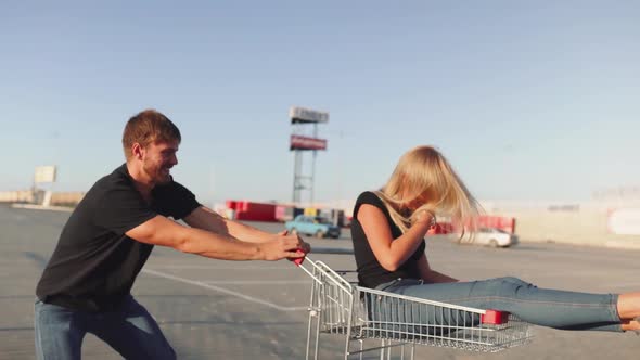 The Guy Rolls Attractive Young Woman in the Trolley on Parking Lot of the Supermarket at Sunset