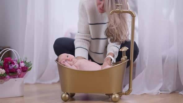 Slim Caucasian Confident Mother Putting Baby Girl in Golden Bath Indoors at Home Talking in Slow