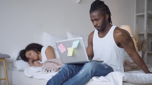 Portrait of Concentrated Serious African American Man Surfing Internet on Laptop Sitting on Bed with