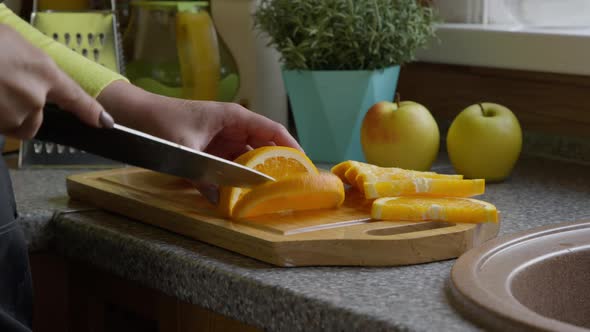Housewife in Apron Cuts an Orange Into Pieces with Kitchen Knife on a Oak Board.
