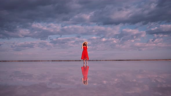 Young Woman in a Waving Dress Walks Along a Pink Lake in Which Clouds Are Reflected.