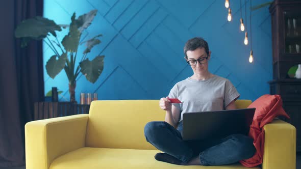 Woman with Short Hair with Glasses Holds a Credit Card While Sitting on the Couch and Enters the