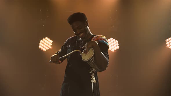 Black Musician Plays Talking African Yuka Drum in a Dark Studio Against a Backdrop of Bright Lights