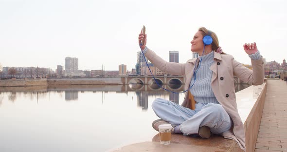 A Young Woman in Headphones and Listens to Relaxing Music on the Embankment