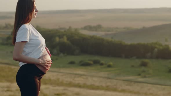 Pregnant Woman is Standing on Hill and Stroking Her Stomach with Two Hands in Circle Performing