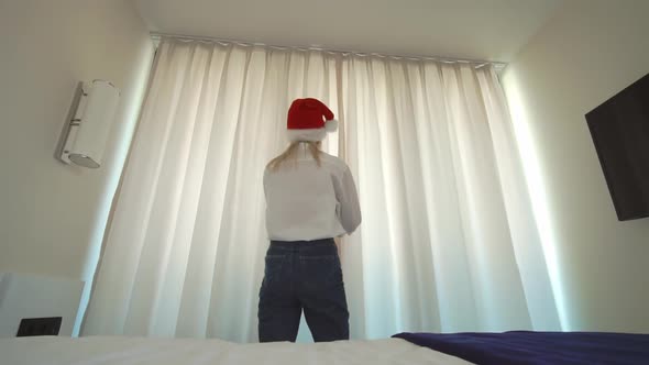 a woman in a Santa Claus hat gets out of bed and opens the window curtains.
