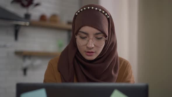 Intelligent Middle Eastern Young Woman in Hijab and Eyeglasses Typing on Laptop Keyboard Messaging