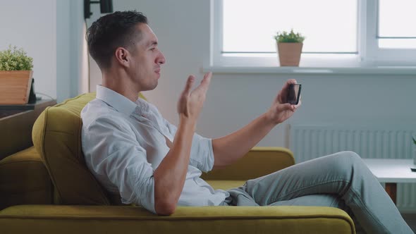 Smiling Young Man with Smartphone Making Video Call While Sitting on Couch in Living Room. Portrait